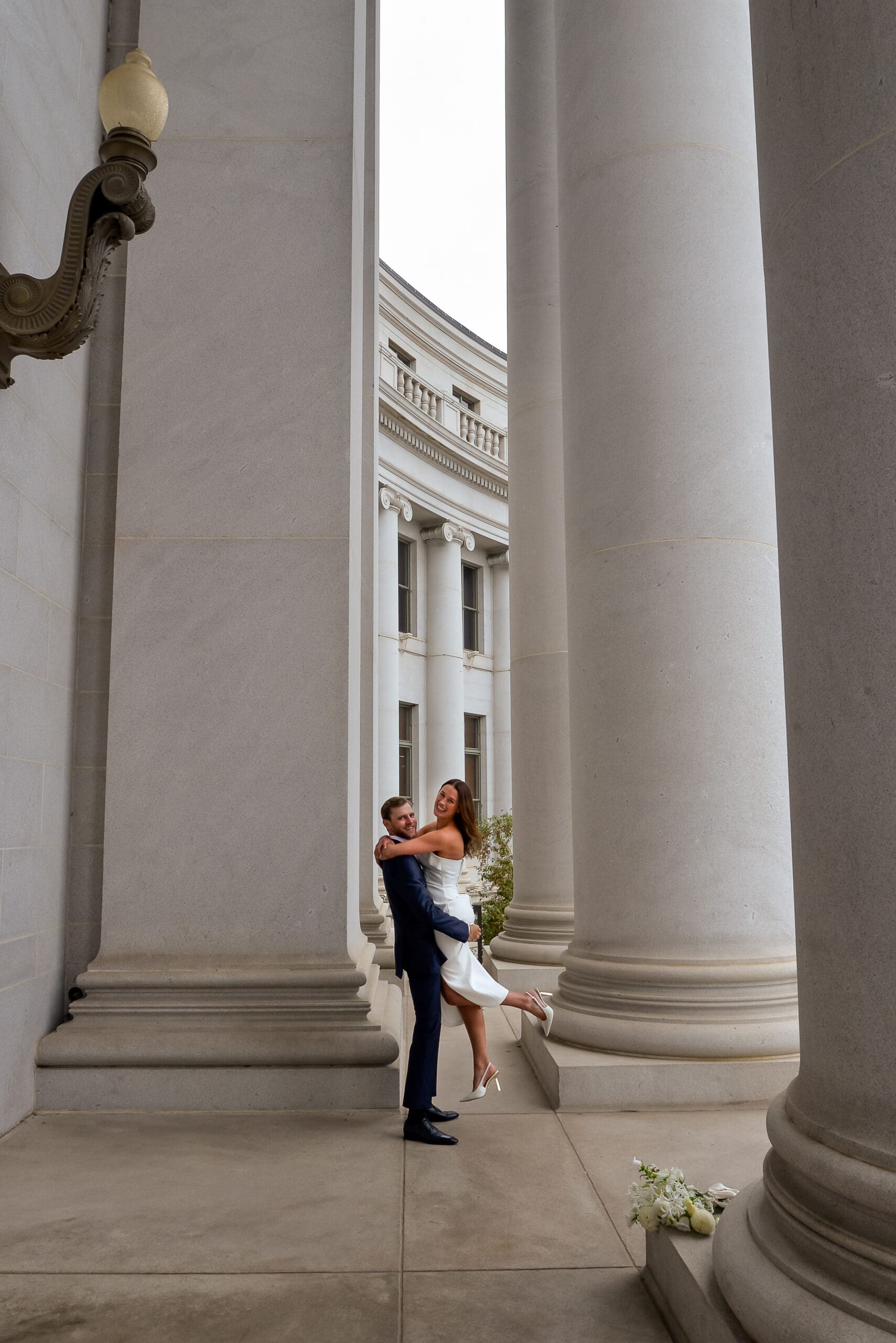 Groom lifts bride up and they both smile at the camera in front of the big white columns at the Denver courthouse