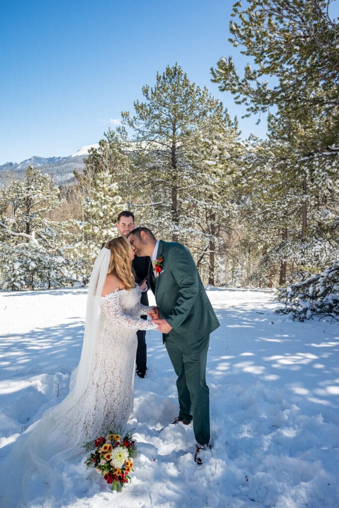 Couple kisses after saying I Do in front of snowy trees and mountains. 