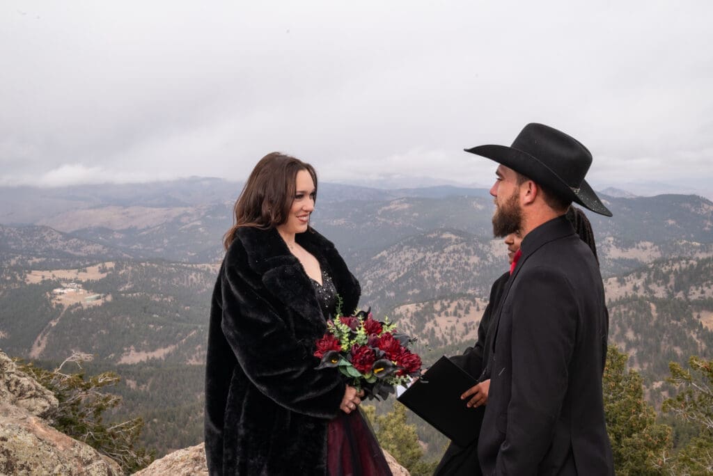 Couple wearing black and red stands on top of Flagstaff Mountain and exchanges vows with an officiant. 