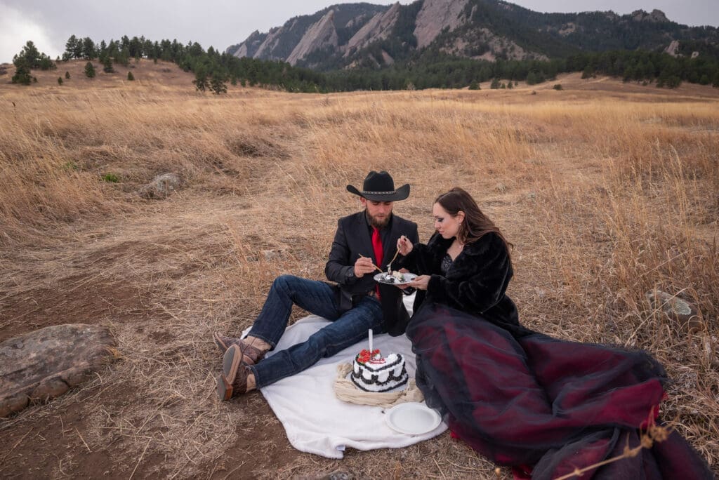 A couple dressed in black and red wedding attire sit on the grass at the base of the Flatirons in Boulder and eat a cake after getting eloped.