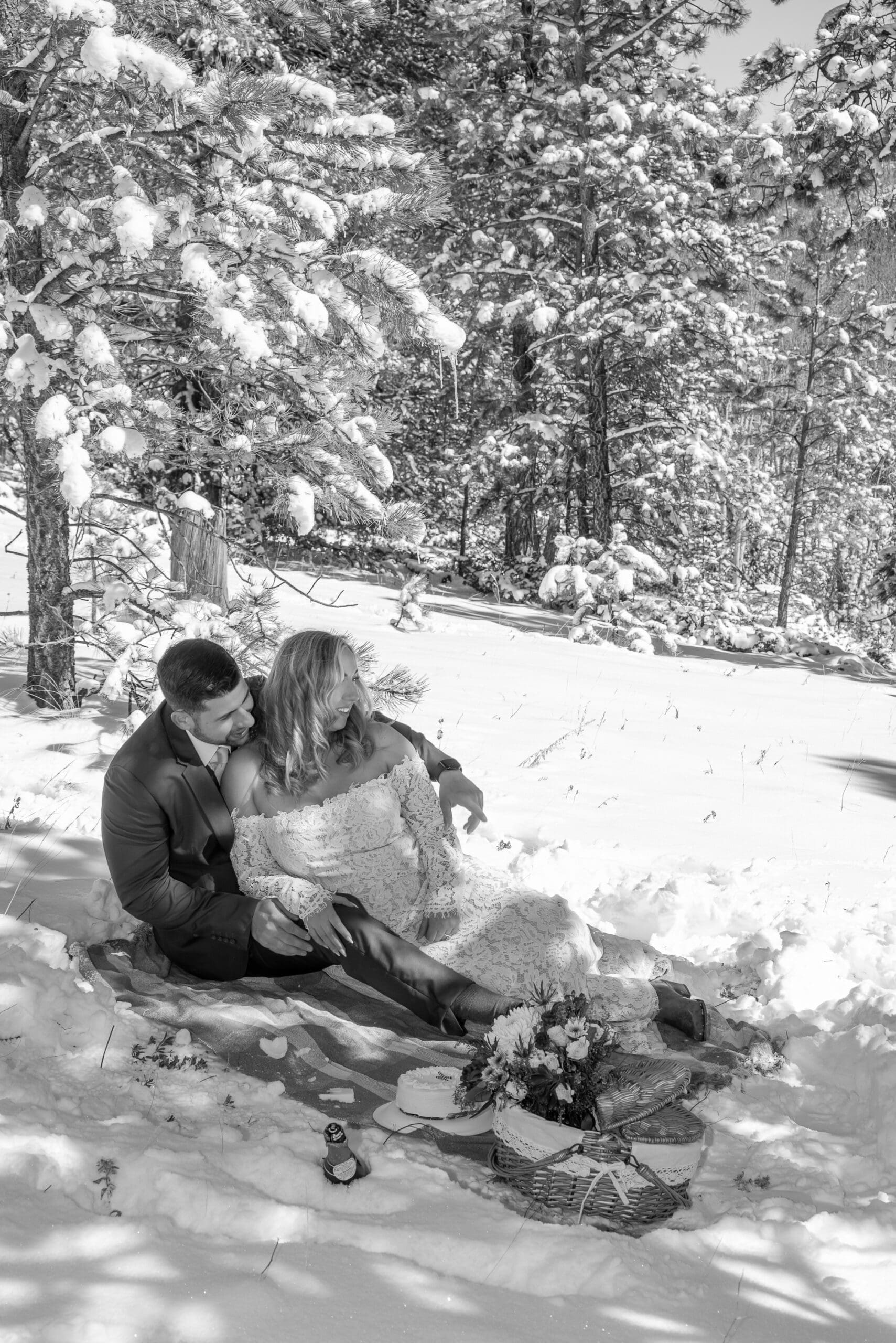 Couple sits on a picnic blanket in the snow, admiring the scenery of pine trees and bright snow in the sunshine.