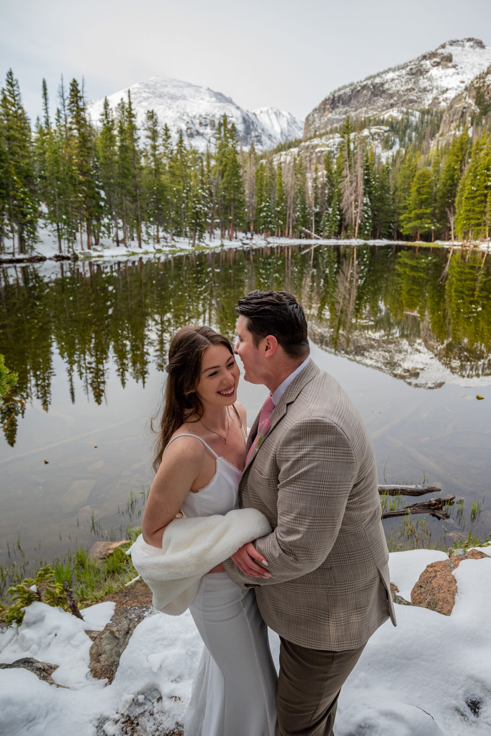 Couple laughs with each other in front of an alpine lake with pine trees and snow covered peaks in the background. What is the Best Time of Year to Elope in Colorado?
